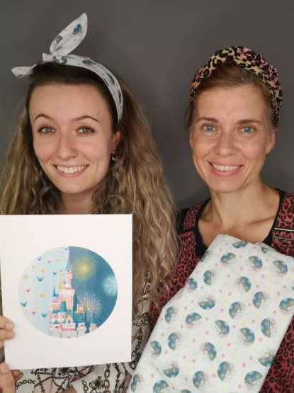 Image of two women, holding up a print inspired by Disney, that the same print as a pattern on fabric. The background is plain grey.