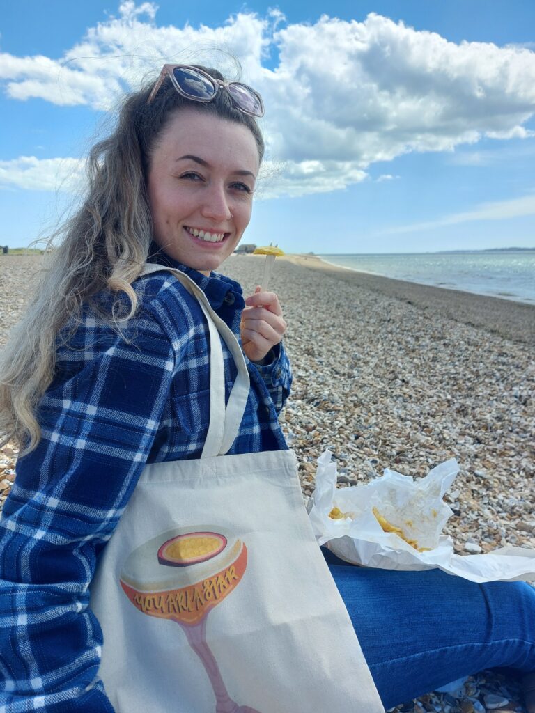 In the background, there is a pebble beach and the sea, with fluffy white clouds in a blue sky. In the foreground, a woman with long, curly brunette hair in her 20s, who is wearing a blue checked shacket, smiles at the camera whilst holding a wooden throwaway fork with a chip on the end. She has a organic cotton tote on her shoulder with a pornstar martini design on the front which says "you are a star" on the glass.