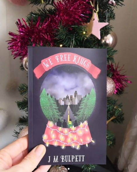 A hand holding a book by the author J M Bulpett - on the front of the cover there is a drawn snowglobe, with four men, a long road leading to a city and trees inside the snowglobe. The title is on a red scarf, and the cover background is navy. Behind the book is a small christmas tree wrapped with pink tinsel and adorned with pink decorations.