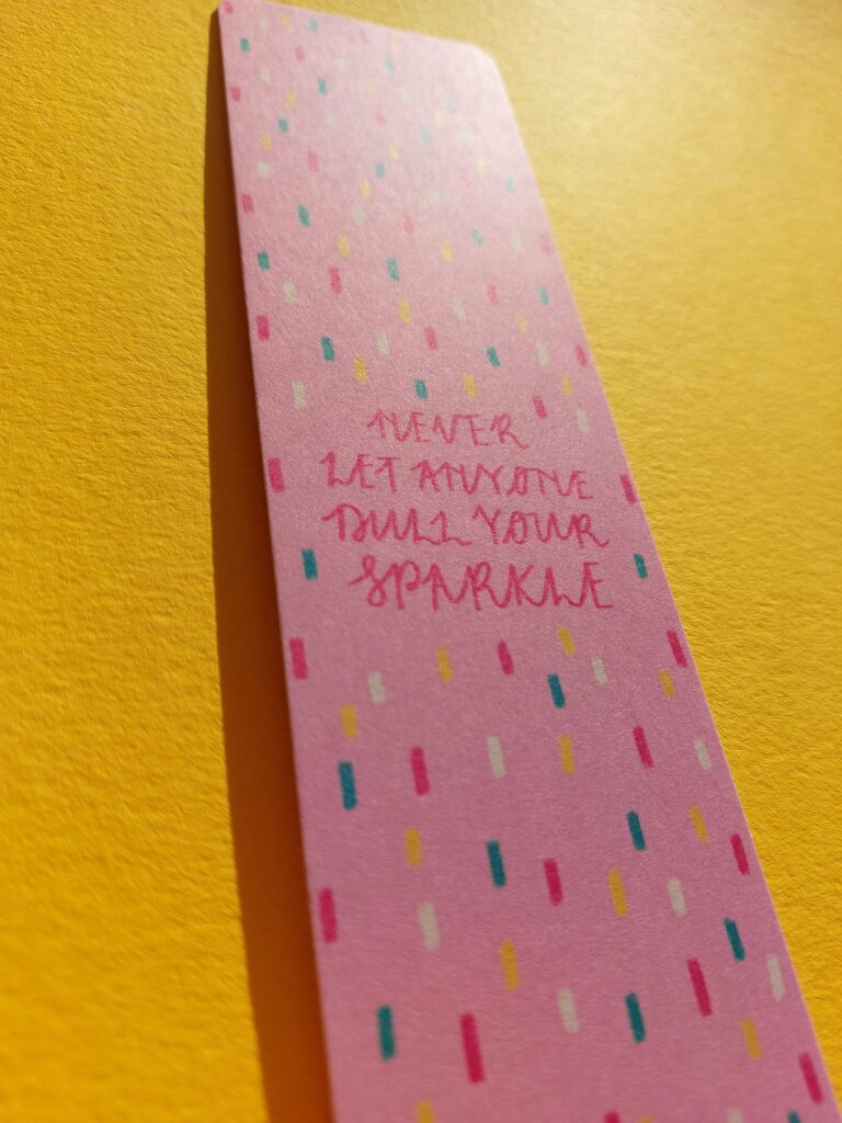 A pink bookmark that reads "never let anyone dull your sparkle" with a multicoloured confetti style pattern in the background. It's sat on a bright, sunny yellow background.