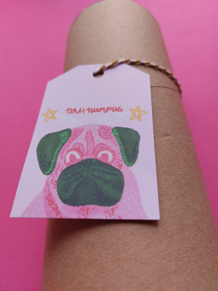 Bah Humpug Christmas Gift Tag, with a pink and green illustrated pug on the gift tag, which has a light pink background, and "bah humpug" in red lettering. It is tied to a cardboard tube with gold and white bakers twine, and is sat on a bright pink background.
