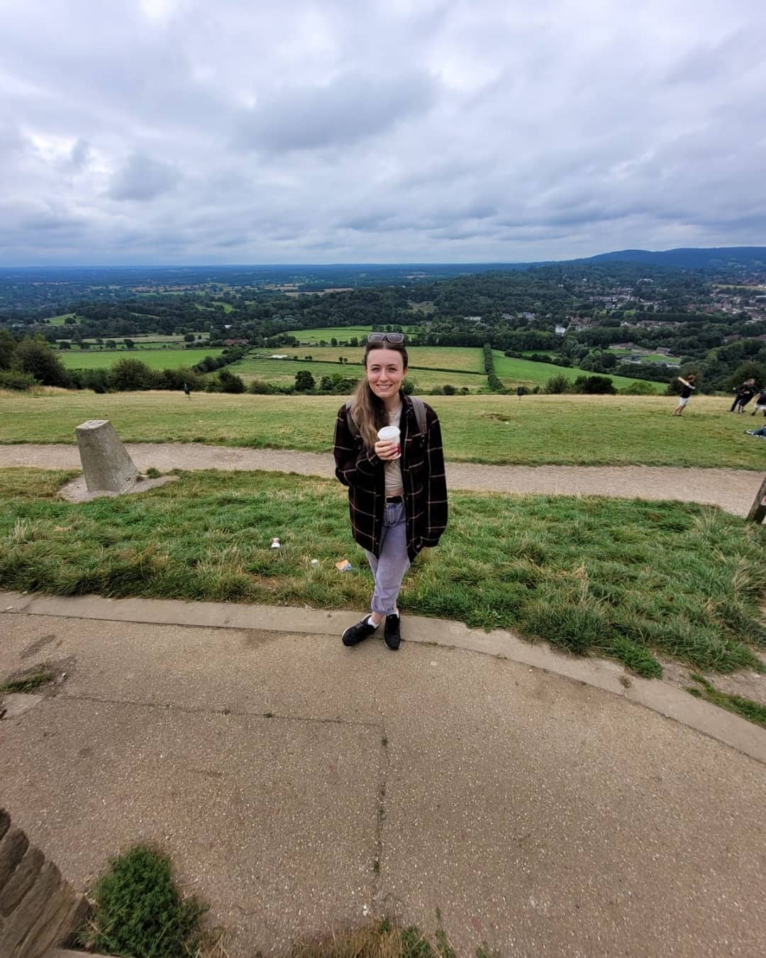 A woman in her twenties with long brown hair, wearing a reddy brown tartan shacket, holding a coffee cup. She (aka me) is standing on top of Box Hill, which is in the Surrey hills, you can see the rolling green hills and the cloudy sky in the background.