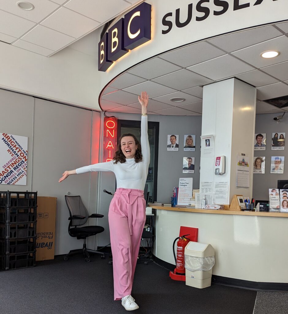 A picture of a brunette woman in her mid twenties wearing Barbie pink trousers and a cream roll neck top. Her arms are extended in the air - one upwards and the other to the left, she is standing in the BBC Sussex Radio reception, it says BBC sussex above her on the wall, and there is an "On Air' red neon sign to the right.