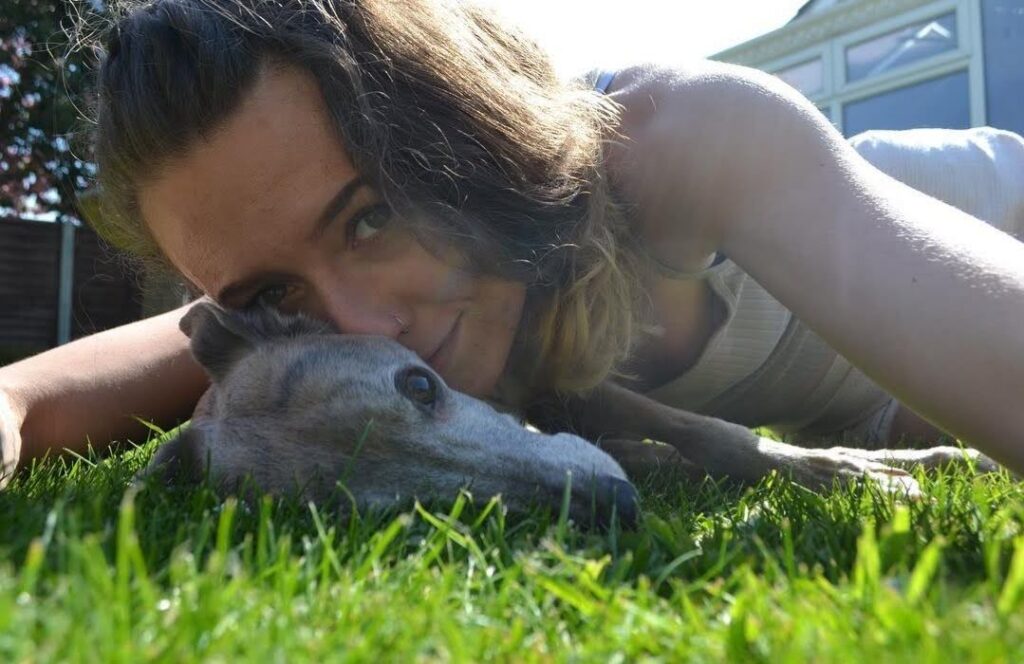 A picture of a young brunette woman laying on her front on the grass with a brindle whippet laying side ways in front of her, both are facing the camera, it's a sunny day!