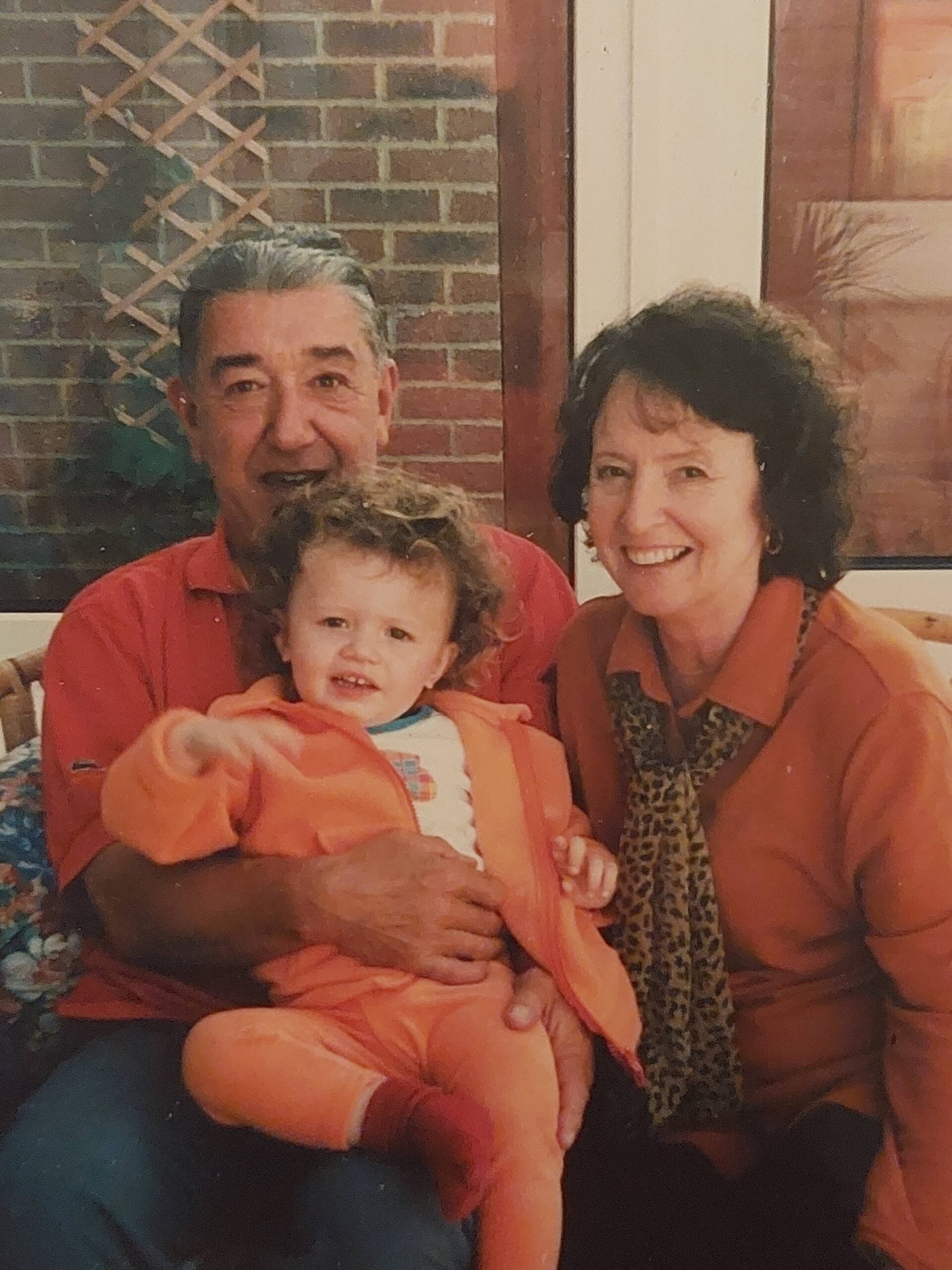 A picture of Hope Bullen with her Grandparents when she was a toddler. She has curly dark blonde hair, and they are all wearing orange. Her Grandad has dark hair and is greying at the front, her Nan has brunette hair, they are sitting in a conservatory.